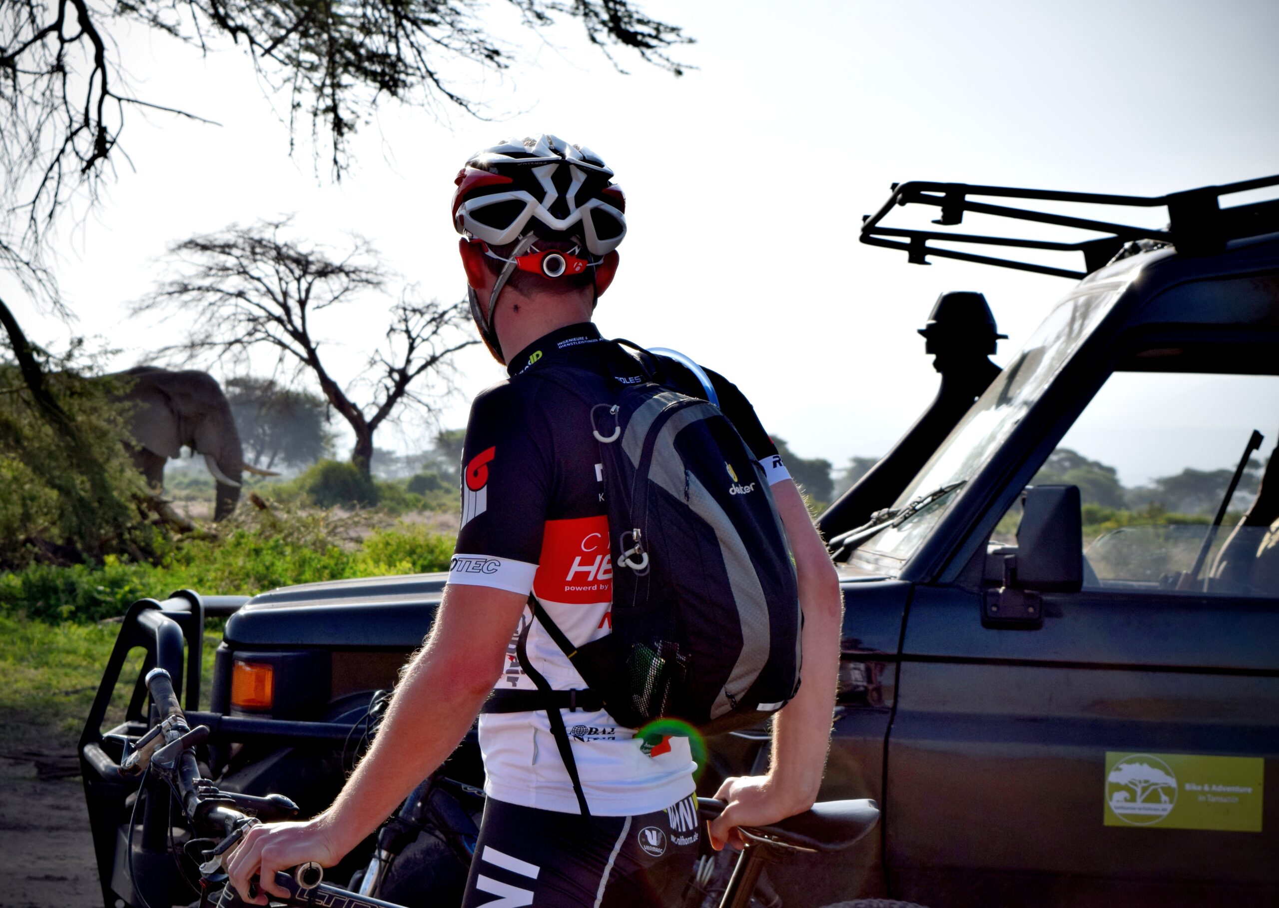 Mountainbiker vor einem Safari-Fahrzeug mit Giraffe im Hintergrund in Tansania