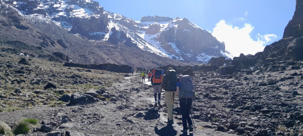 Trekkerguppe auf gerölligem Pfad unterhalb des schneebedeckten Kilimanjaro-Gipfels