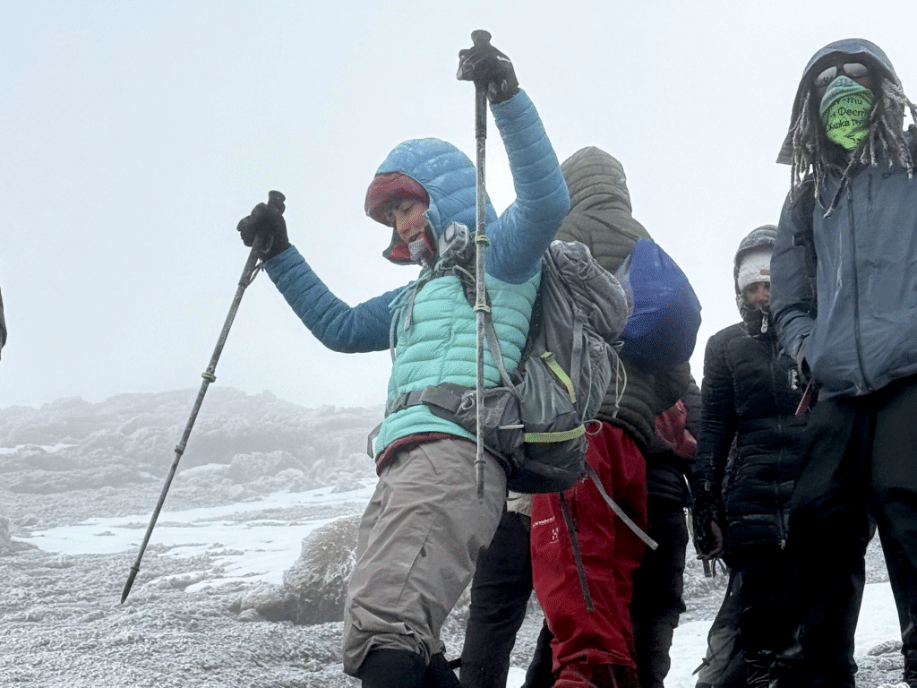Frau steht stolz am Gipfel des Kilimanjaro und reißt ihre Wanderstöcke in die Höhe, umgeben von Nebel und Eis