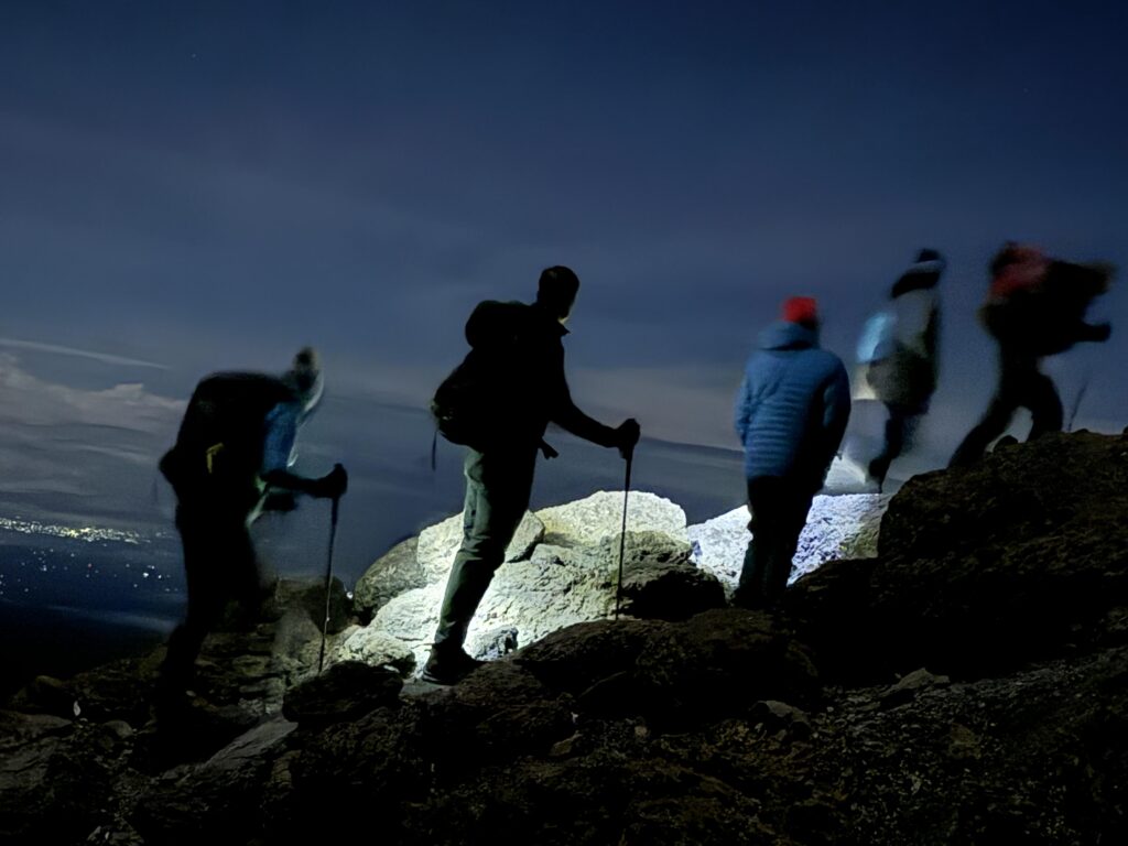 Wanderer mit Stirnlampen am Kilimanjaro blicken im Dunkeln auf die leuchtenden Lichter der Stadt im Tal