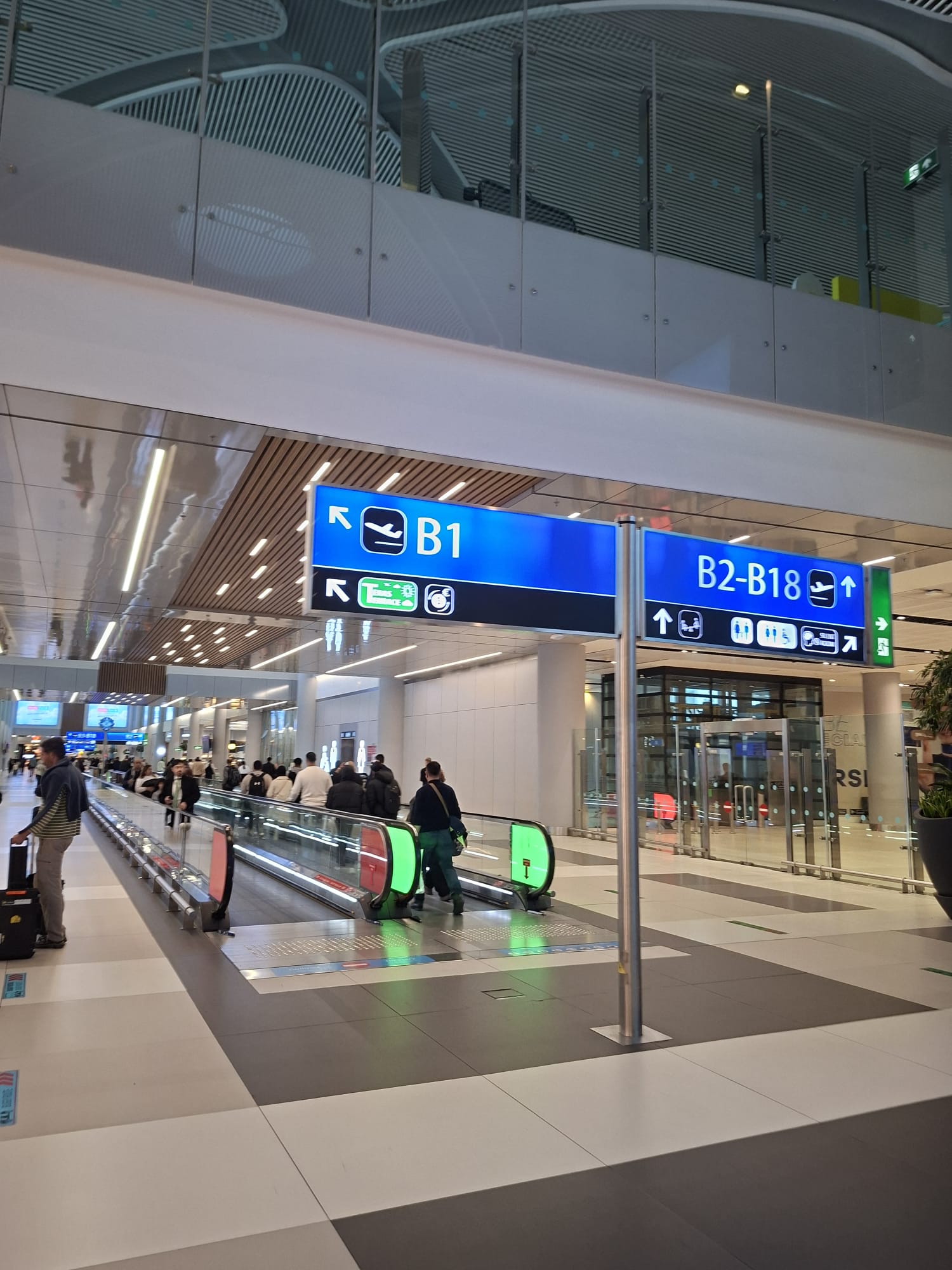 Reisende auf einem Fahrsteig im Istanbul Airport unter Wegweisern zu den Abfluggates