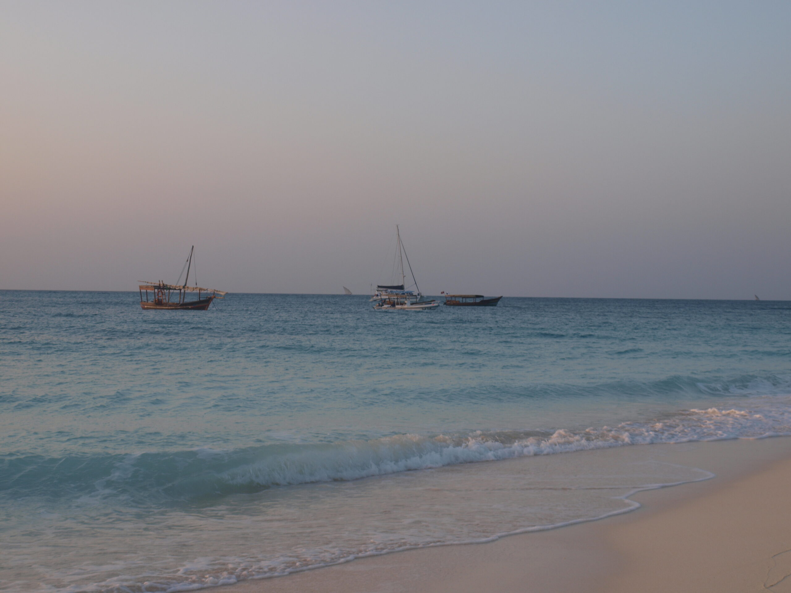 Mehrere Boote vor der Küste von Sansibar und kleine Wellen an einem Sandstrand.